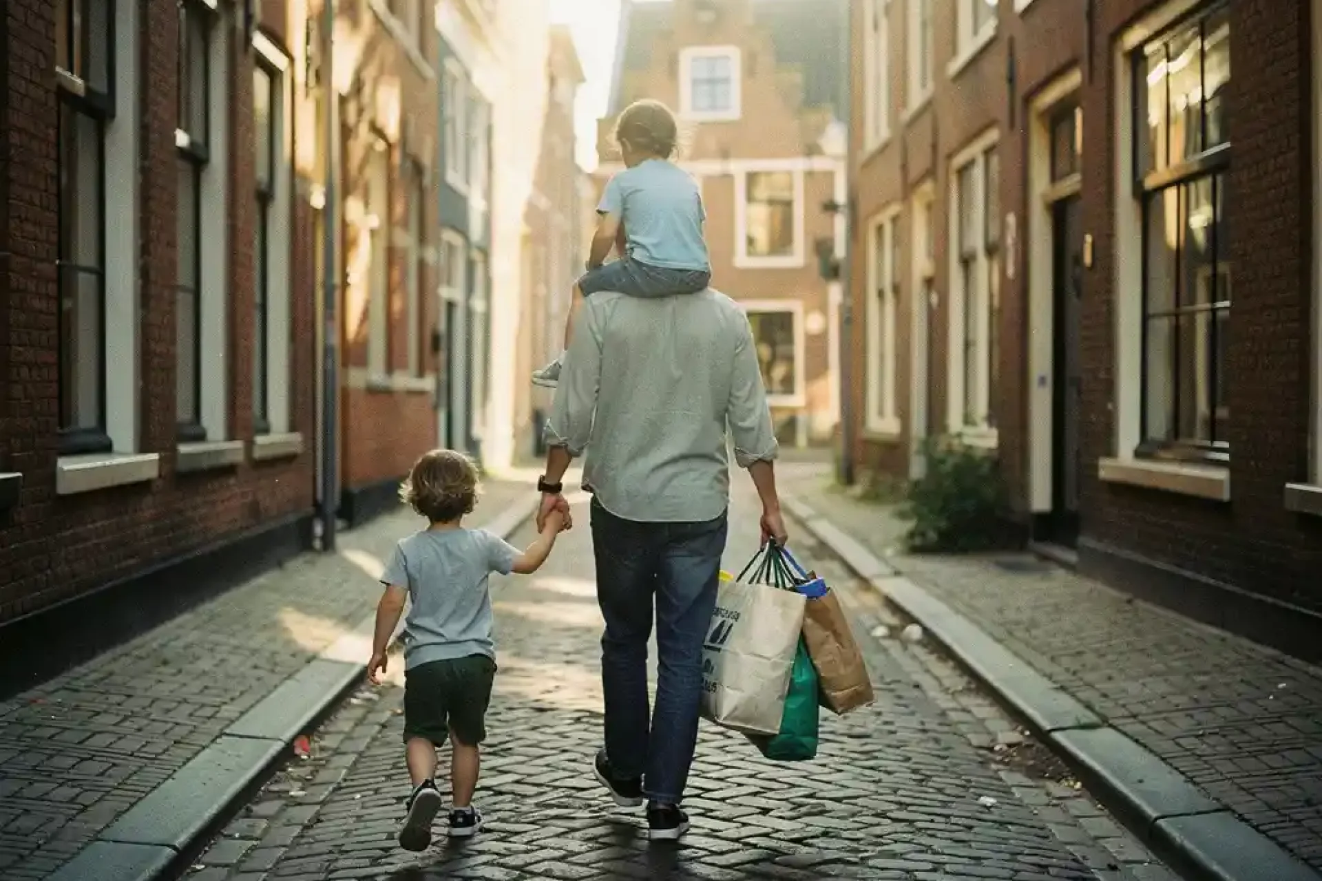 Ouder draagt peuter op schouders terwijl ze boodschappentassen vasthoudt in smalle Hollandse straat, ander kind loopt mee