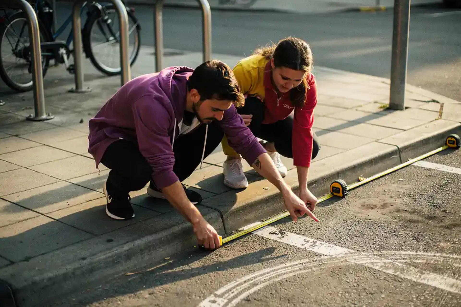 Twee personen meten met meetlint een smalle parkeerplaats in de stad, omringd door fietsrekken en stoeptegels
