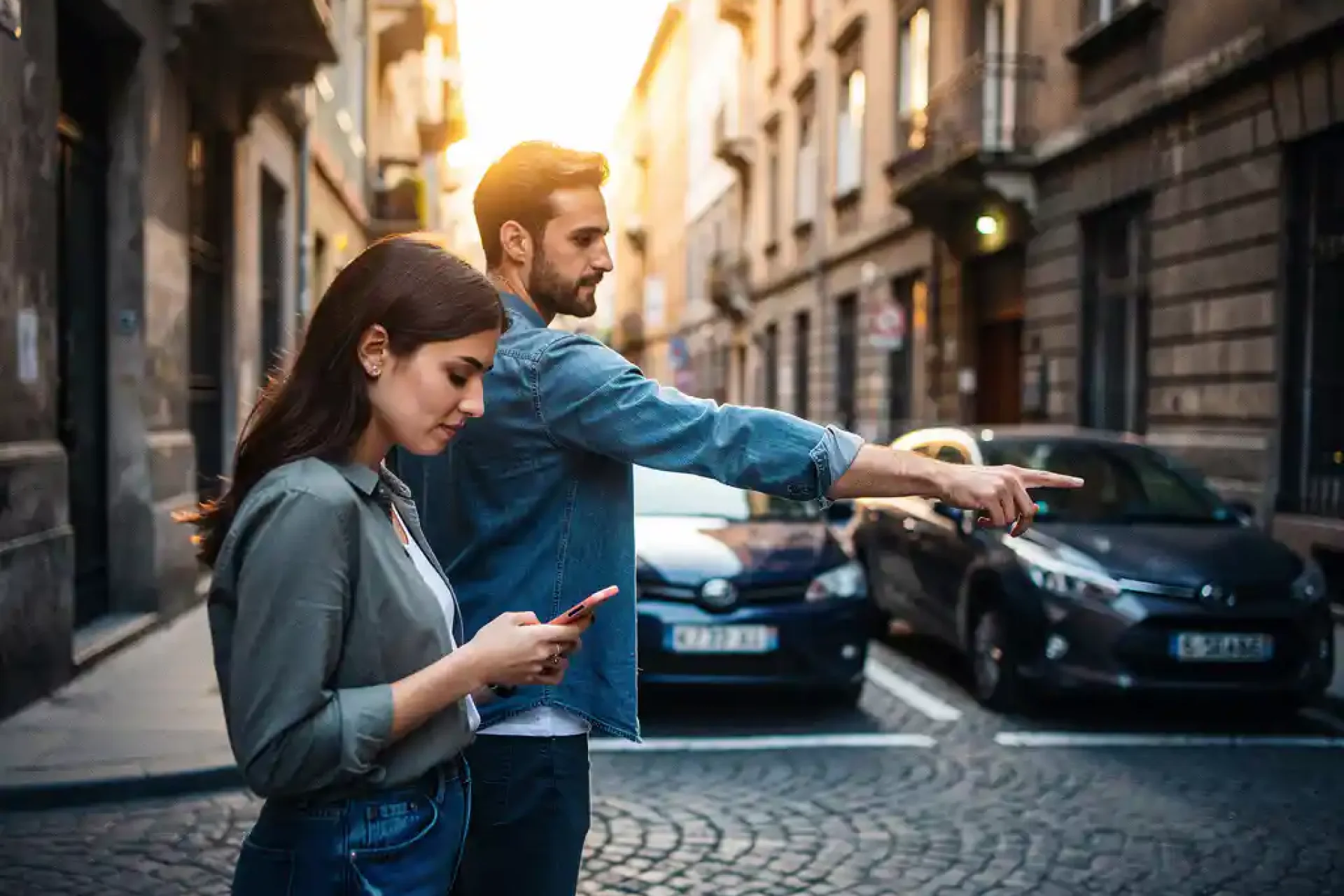 Jong stel op Europese kasseienstraat, vrouw bekijkt smartphone terwijl man naar smalle parkeerplaats wijst tussen geparkeerde auto's