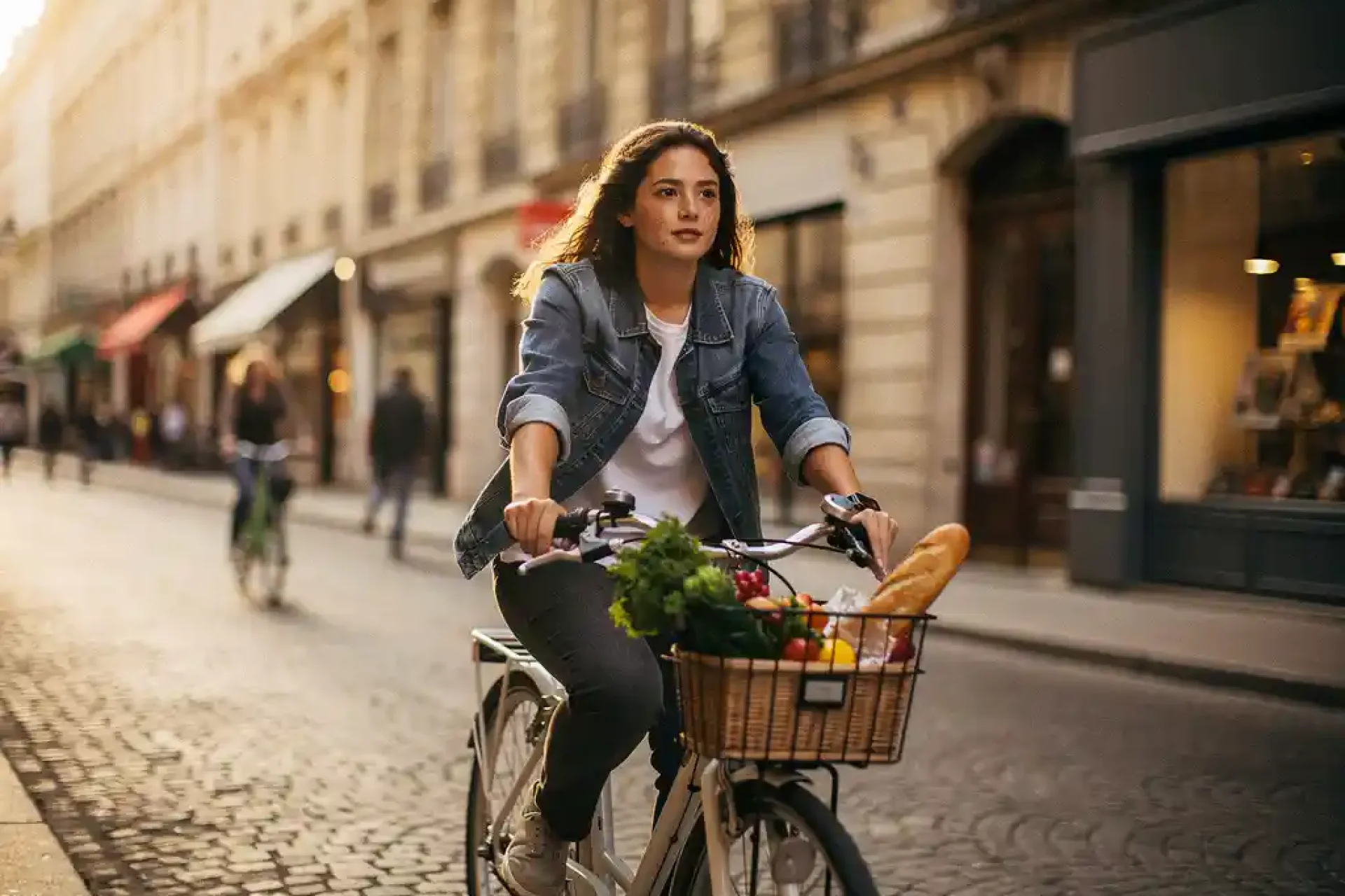Jonge vrouw fietst zelfverzekerd door Europese binnenstad met boodschappen in fietsmand, gouden middaglicht op kasseienstraat.