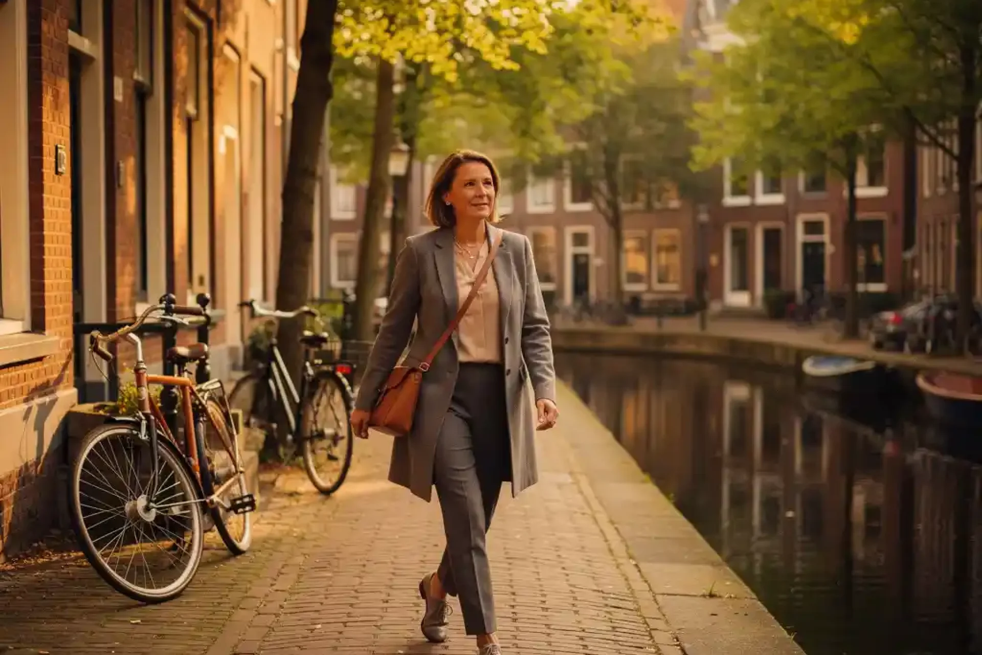 Vrouw van middelbare leeftijd loopt langs een smalle Amsterdamse gracht met bakstenen huizen, fietsen en kinderkopjes in warm middaglicht.