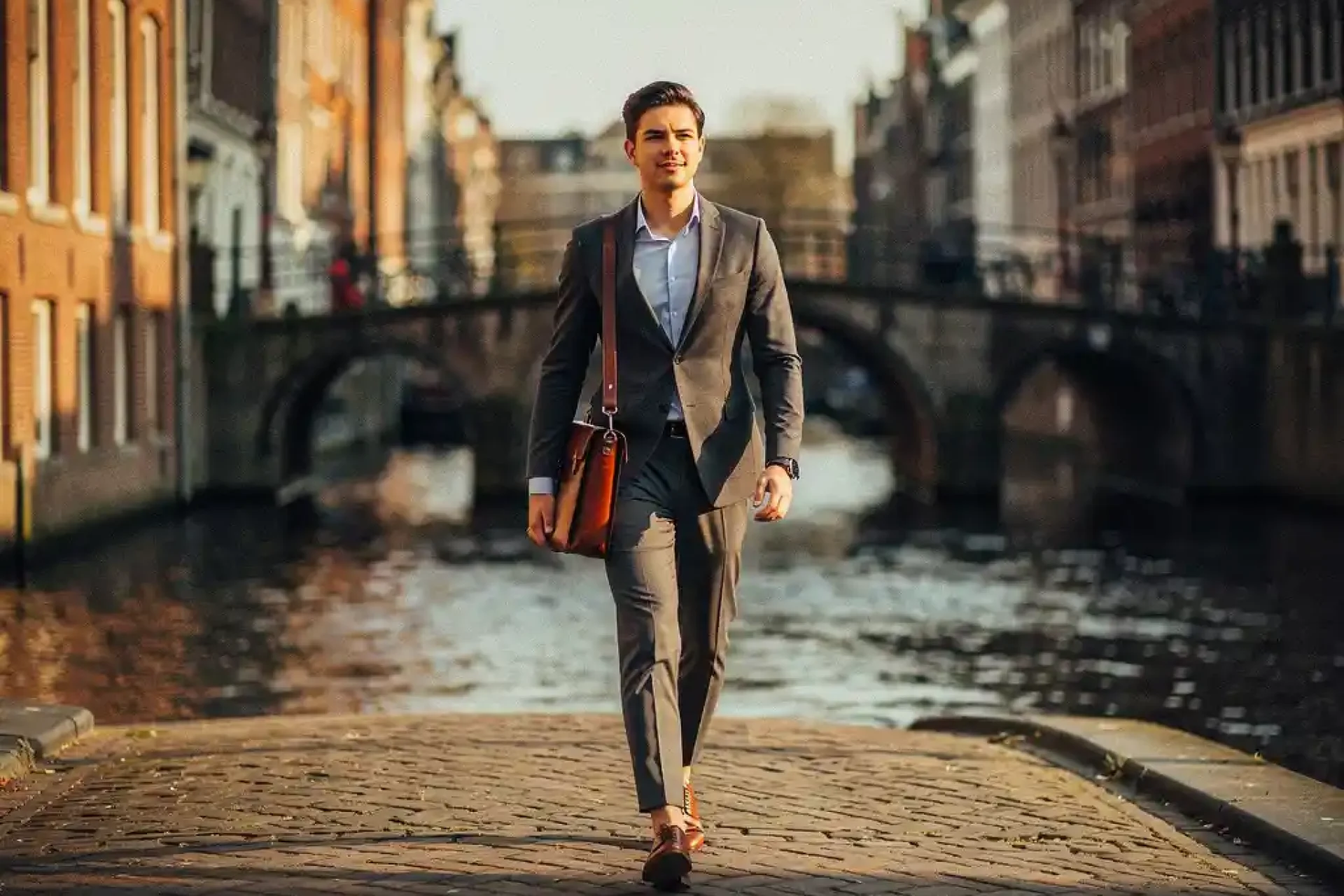 Jonge professional met leren laptoptas loopt zelfverzekerd over een Amsterdamse keistraat langs grachten en boogbruggen in gouden middaglicht.