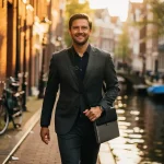 Professional met laptoptas wandelt door smalle Amsterdamse gracht, gouden middaglicht op historische gevels en water.