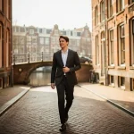 Jonge professional in getailleerd jasje loopt zelfverzekerd door smalle Amsterdamse gracht met kinderkopjes en bakstenen gevels.