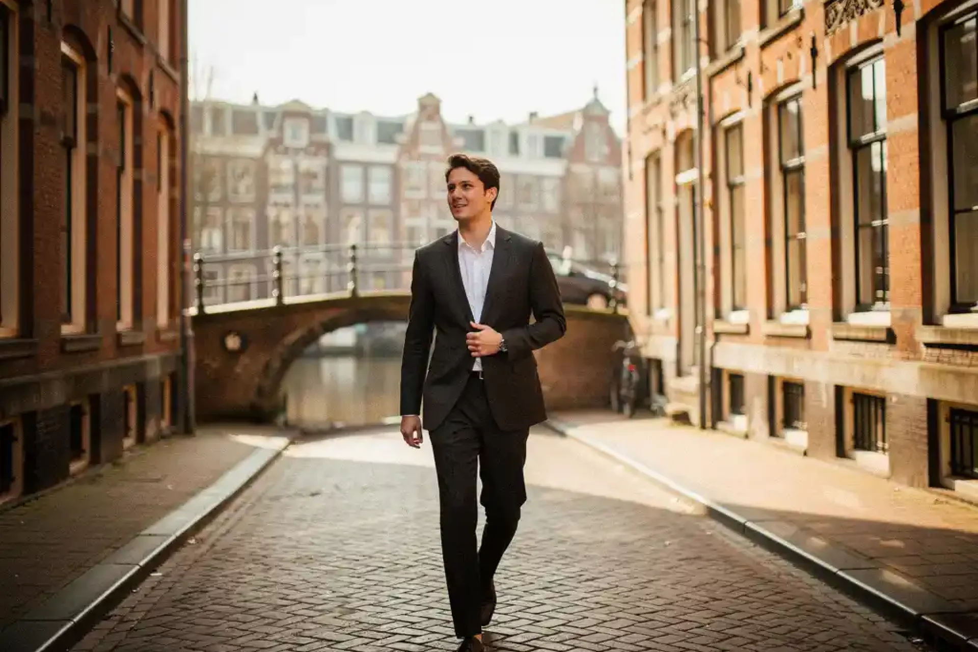 Jonge professional in getailleerd jasje loopt zelfverzekerd door smalle Amsterdamse gracht met kinderkopjes en bakstenen gevels.