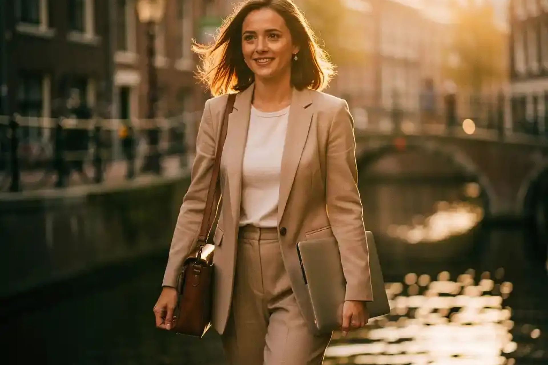 Jonge vrouw met laptoptas wandelt zelfverzekerd door smalle Amsterdamse gracht in gouden ochtendlicht.