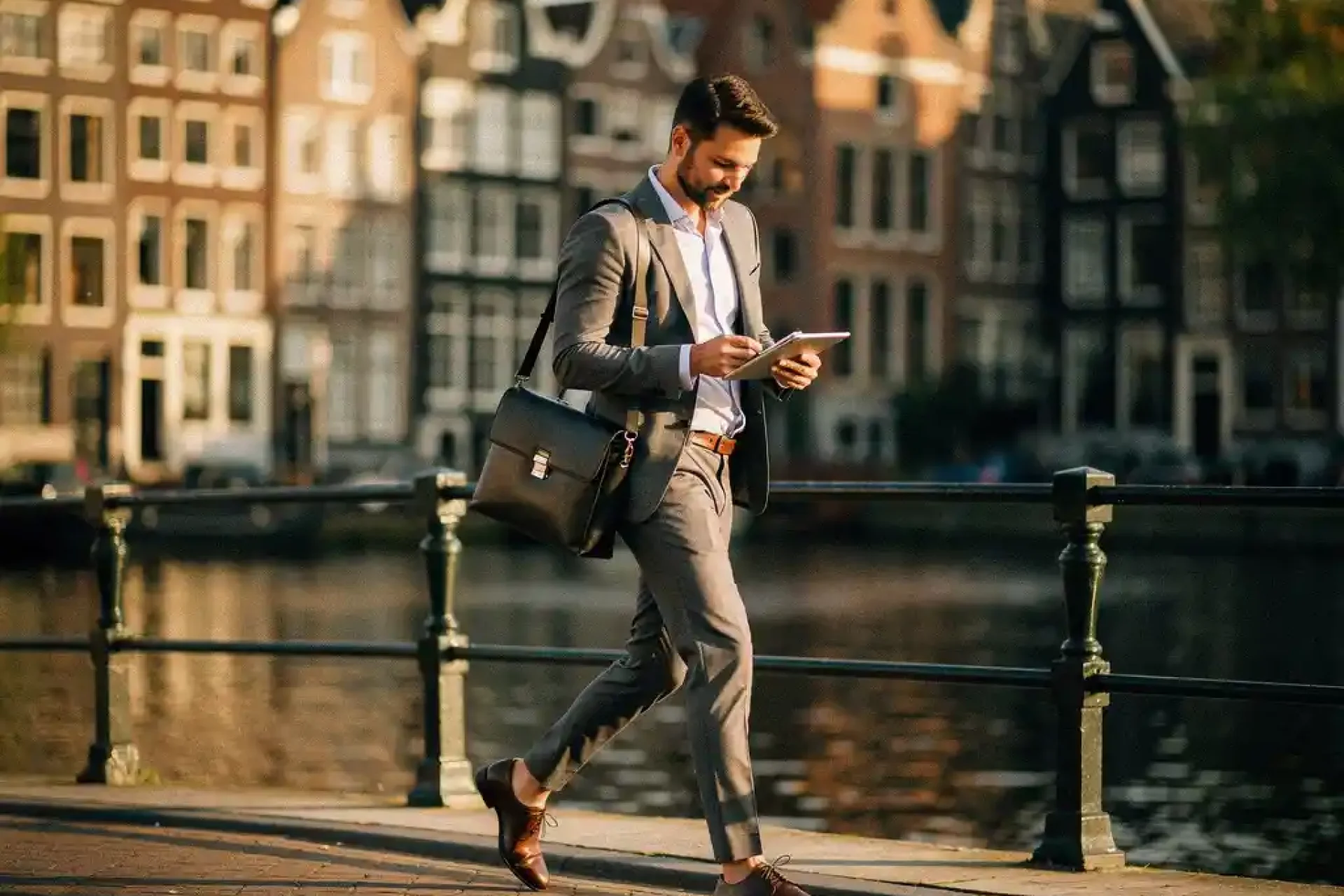 Consultant met laptoptas loopt zelfverzekerd over een Amsterdamse grachtenbrug, smalle grachtenpanden en waterreflecties op de achtergrond.