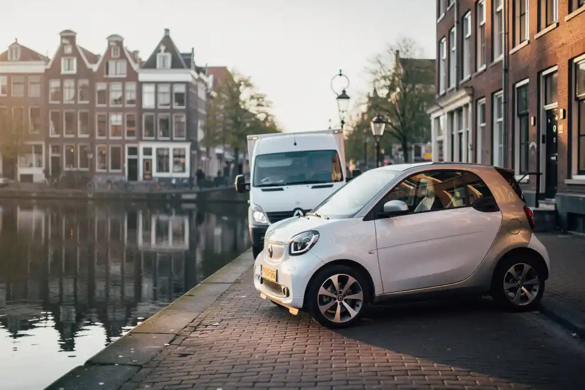 Compacte elektrische stadsauto geparkeerd langs een Amsterdamse gracht, met grachtenpanden weerspiegeld in het water en kinderkopjes op de voorgrond.