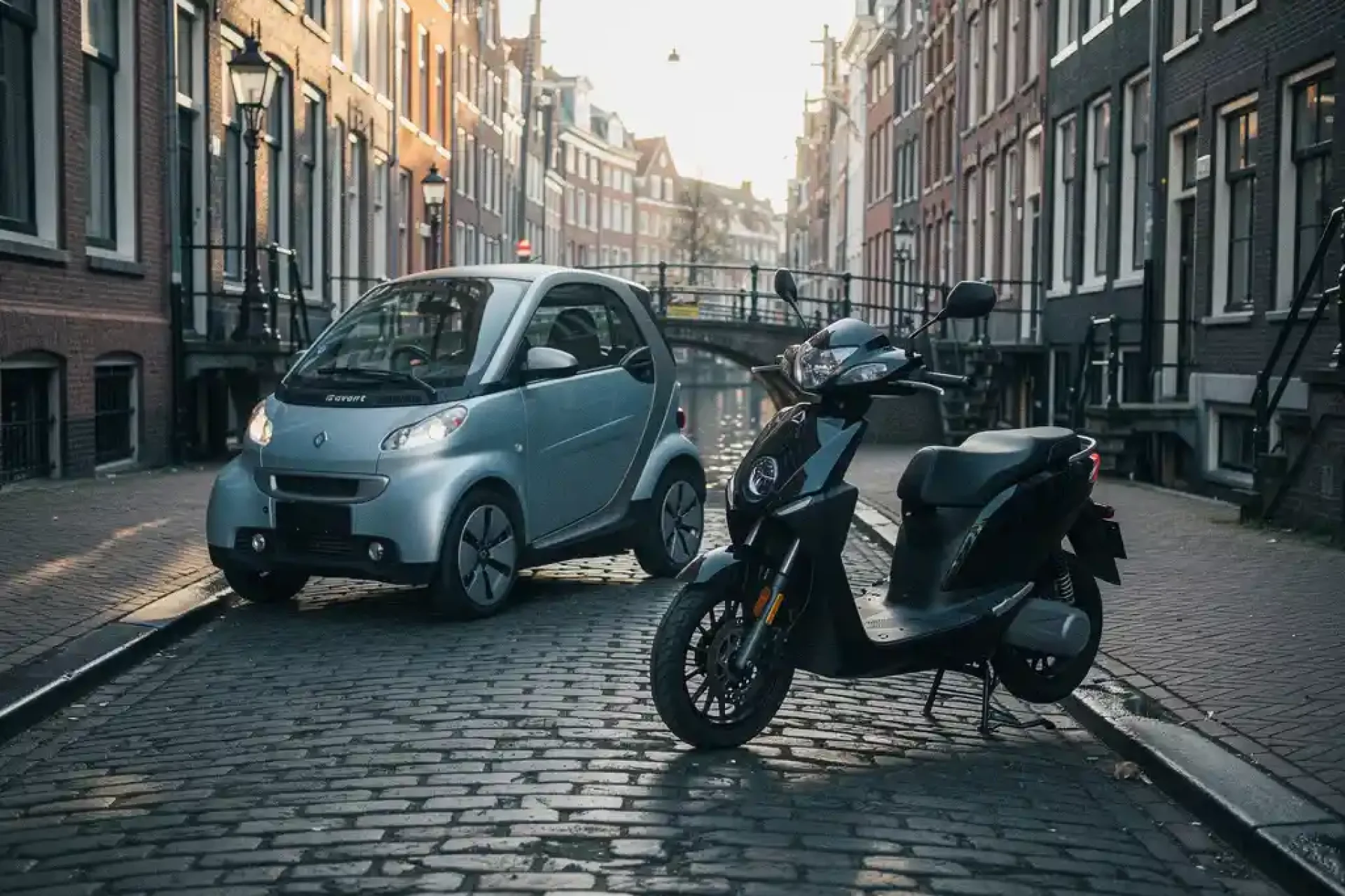 Elektrische stadsauto en scooter op Amsterdamse keistraat langs gracht, historische gevels en brug op achtergrond.