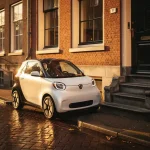 Kleine witte elektrische stadsauto geparkeerd in smalle Amsterdamse straat naast traditionele bakstenen gevel, gouden middaglicht op kinderkopjes.