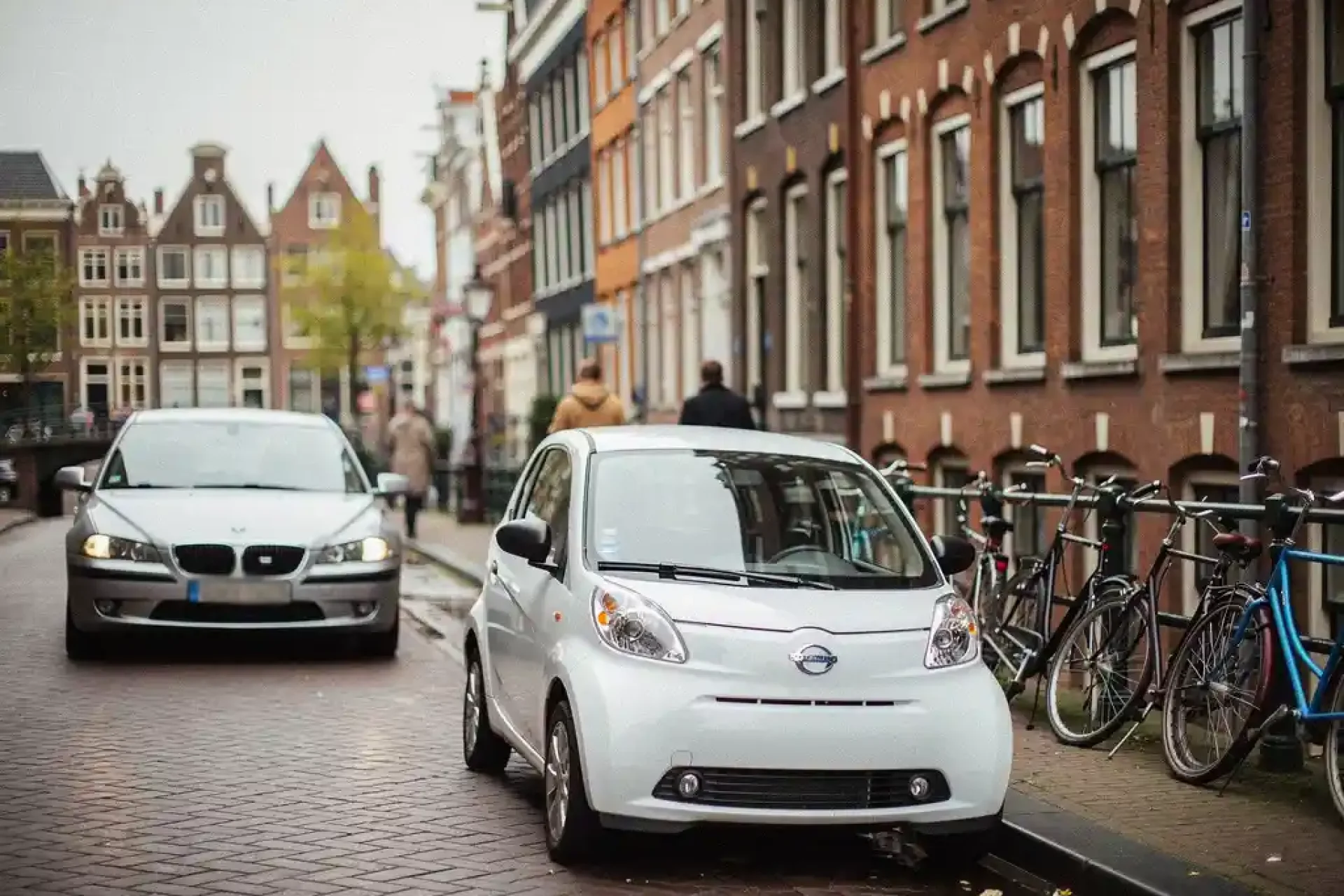 Kleine witte elektrische stadsauto parkeert in smalle Amsterdamse straat langs gracht, grote sedan rijdt rondjes op keienweg.