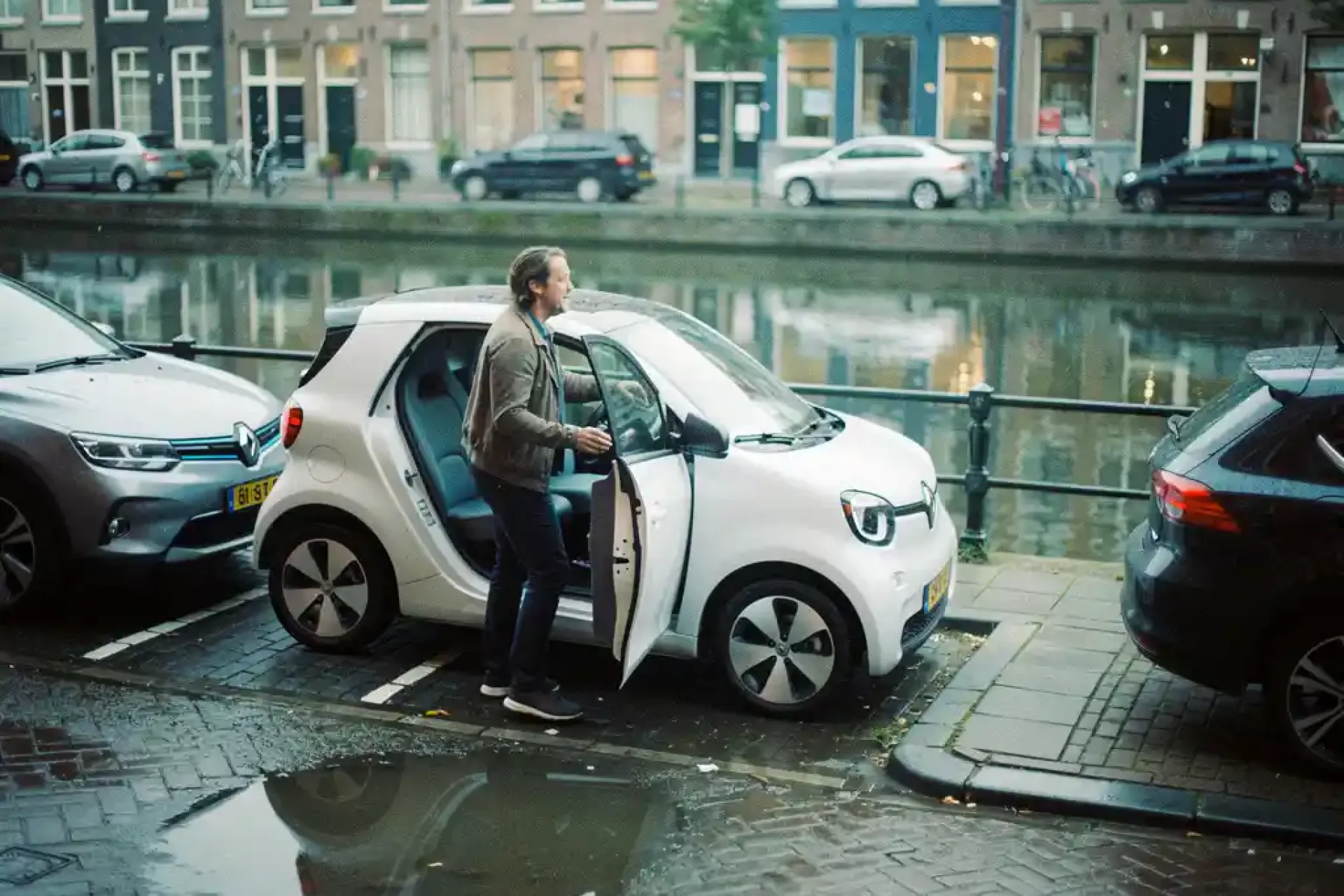 Compacte elektrische stadsauto geparkeerd in smalle Rotterdamse straat, bestuurder stapt uit op keienpad met grachtenreflecties op de achtergrond.
