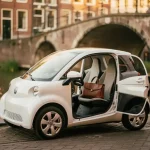 Witte elektrische microcar geparkeerd op een Amsterdamse grachtenstraat, met een leren laptoptas op de passagiersstoel en historische gevels op de achtergrond.
