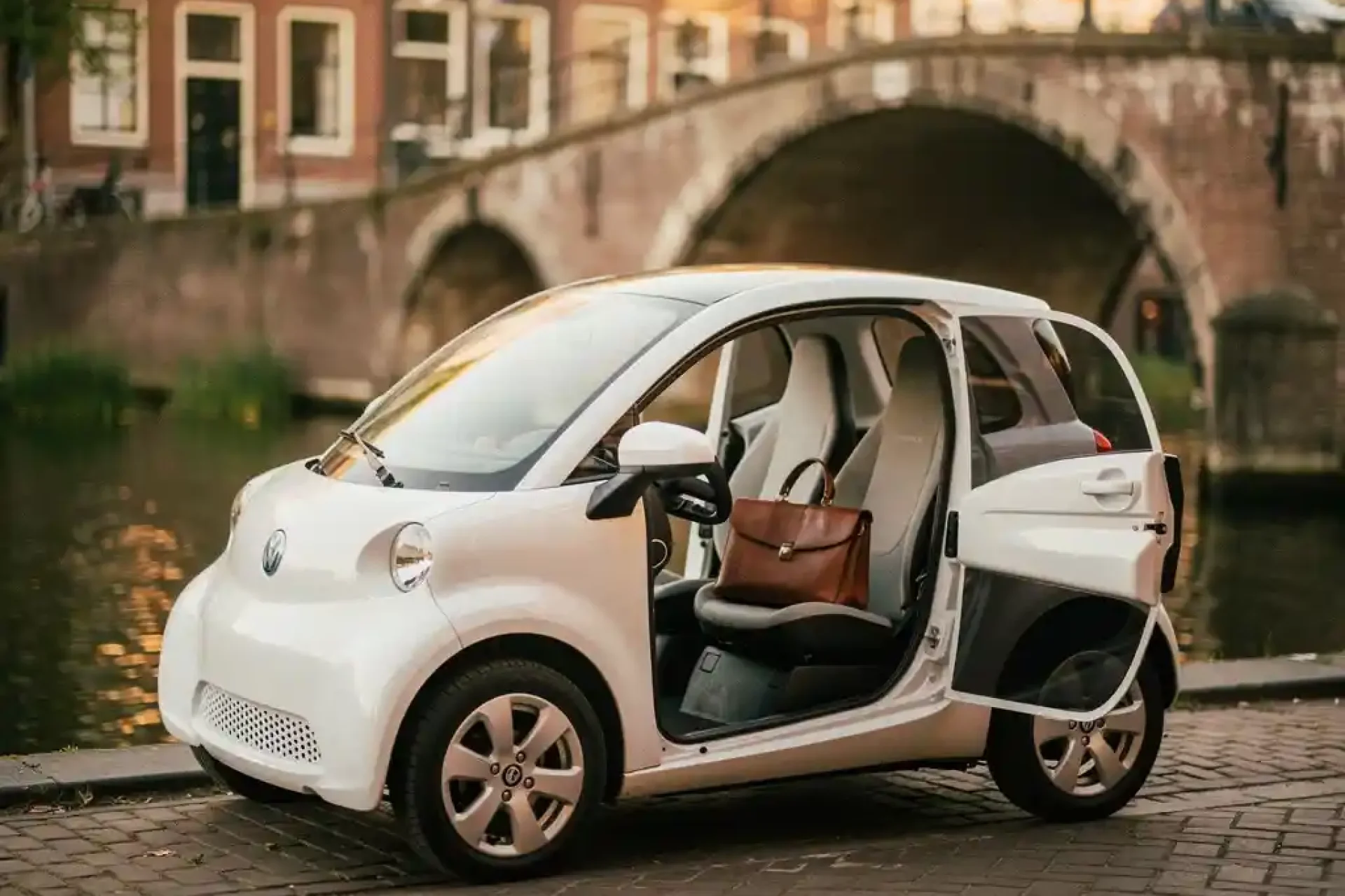 Witte elektrische microcar geparkeerd op een Amsterdamse grachtenstraat, met een leren laptoptas op de passagiersstoel en historische gevels op de achtergrond.