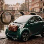 Compacte elektrische microcar geparkeerd langs een Amsterdams gracht, lederen tas op de motorkap, smalle gevels en boogbrug op de achtergrond.