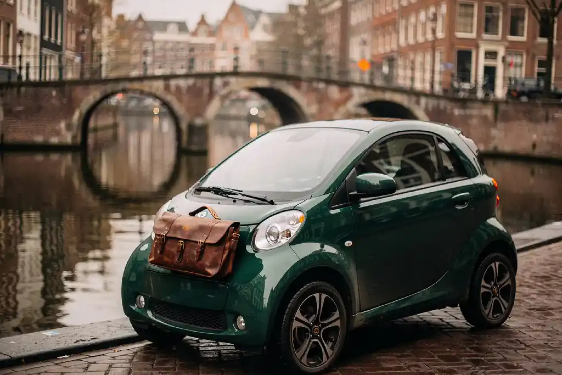Compacte elektrische microcar geparkeerd langs een Amsterdams gracht, lederen tas op de motorkap, smalle gevels en boogbrug op de achtergrond.