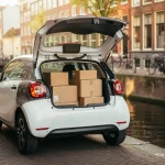 Elektrische stadsauto met open achterklep vol bezorgpakketten langs Amsterdams kanaal met fietsen en bakstenen gevels.