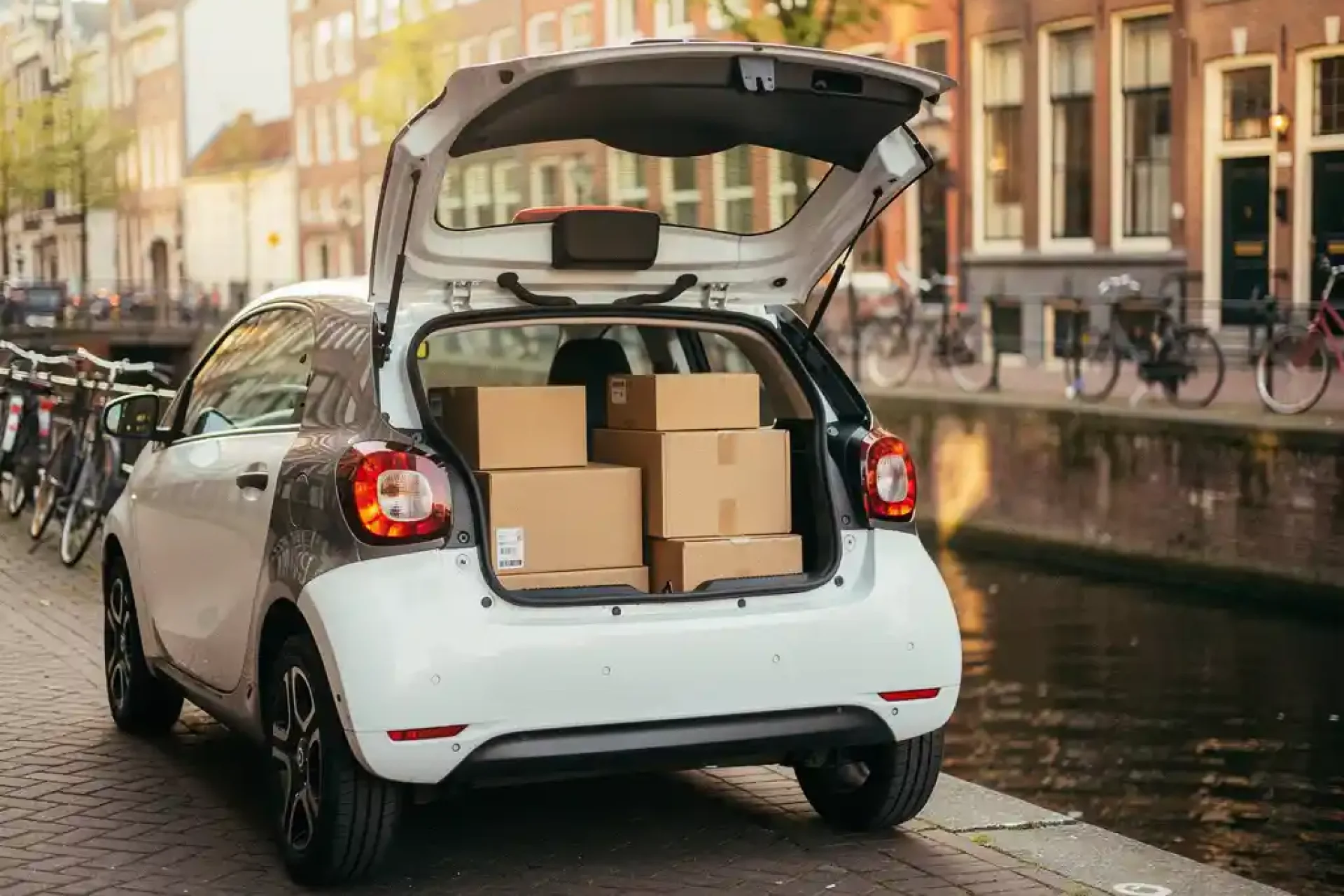 Elektrische stadsauto met open achterklep vol bezorgpakketten langs Amsterdams kanaal met fietsen en bakstenen gevels.
