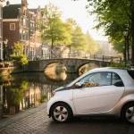 Compacte elektrische stadsauto geparkeerd op smalle Amsterdamse gracht, historische gevels en boogbrug weerspiegeld in het water, warm ochtendlicht.