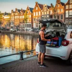 Personal trainer laadt sportuitrusting in witte elektrische stadsauto naast Amsterdams gracht, historische gevels op achtergrond.