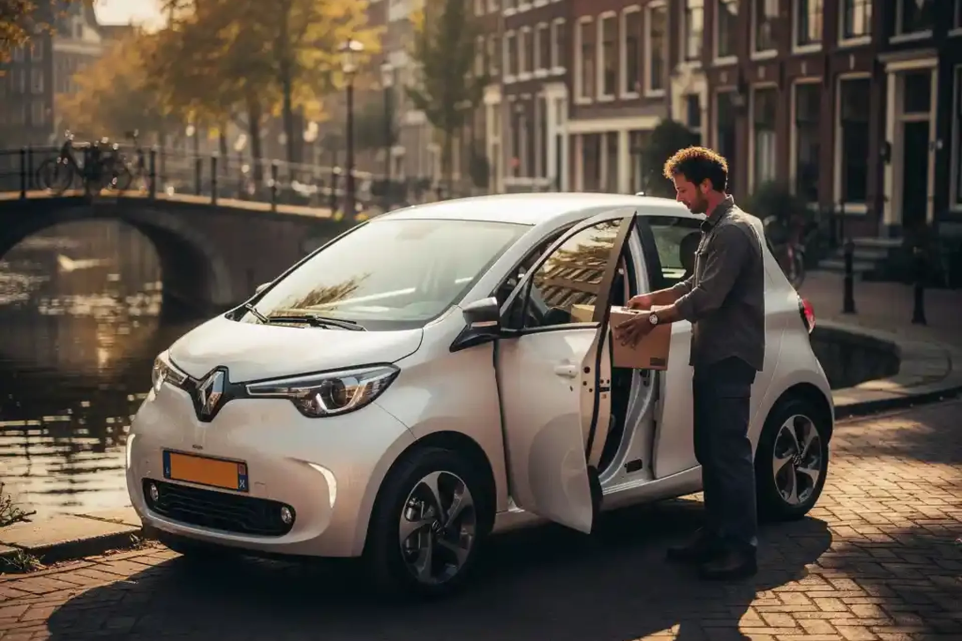 Shopkeeper laadt bezorgdoos in compacte elektrische stadsauto op smalle Amsterdamse gracht, bakstenen grachtenpanden op achtergrond.