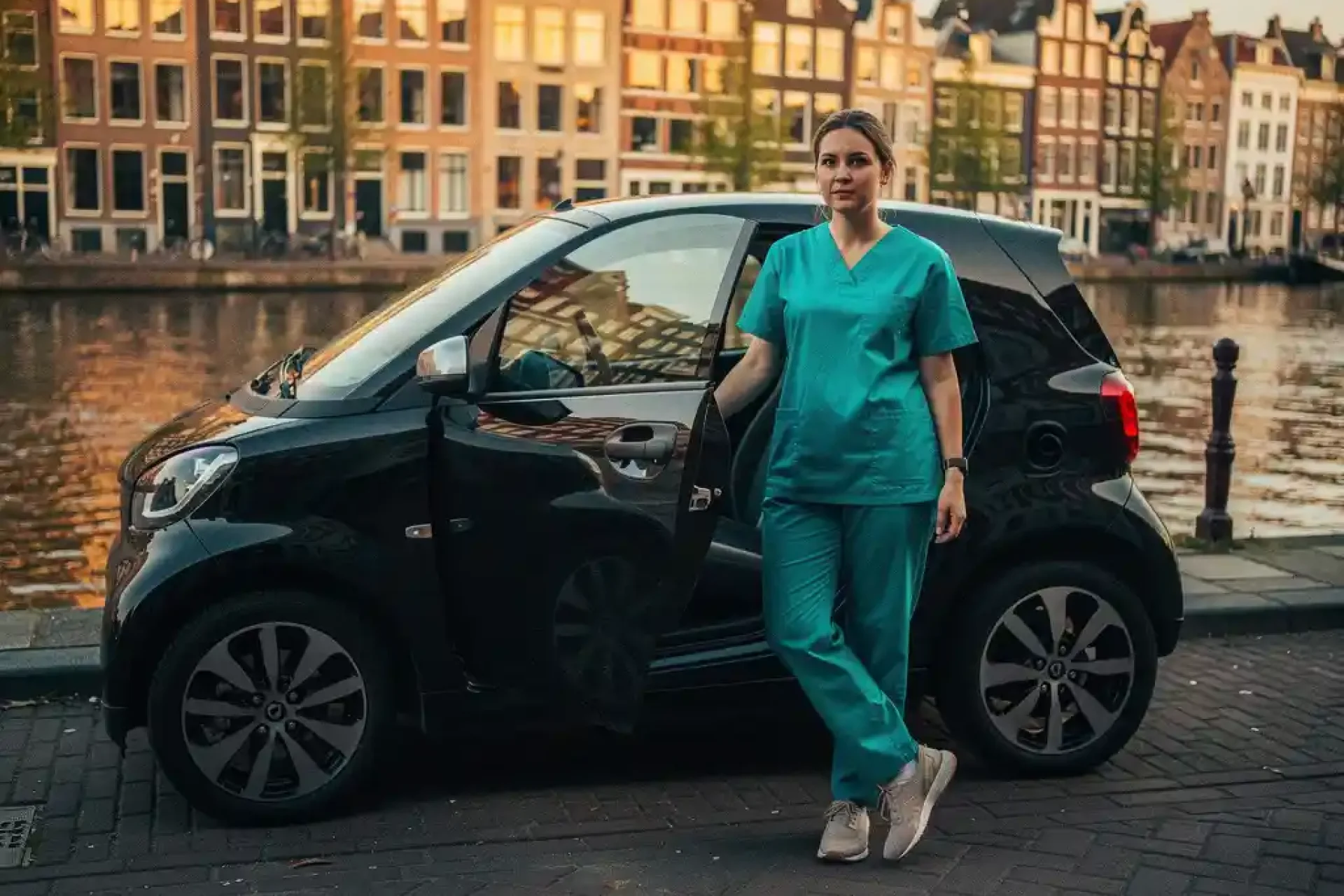 Verpleegkundige stapt uit elektrische stadsauto naast Amsterdams gracht met weerspiegeling van grachtenpanden in autoruit.