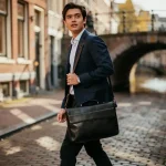 Jonge professional in getailleerd jasje met leren laptoptas loopt zelfverzekerd door smalle Amsterdamse grachtenstraat.
