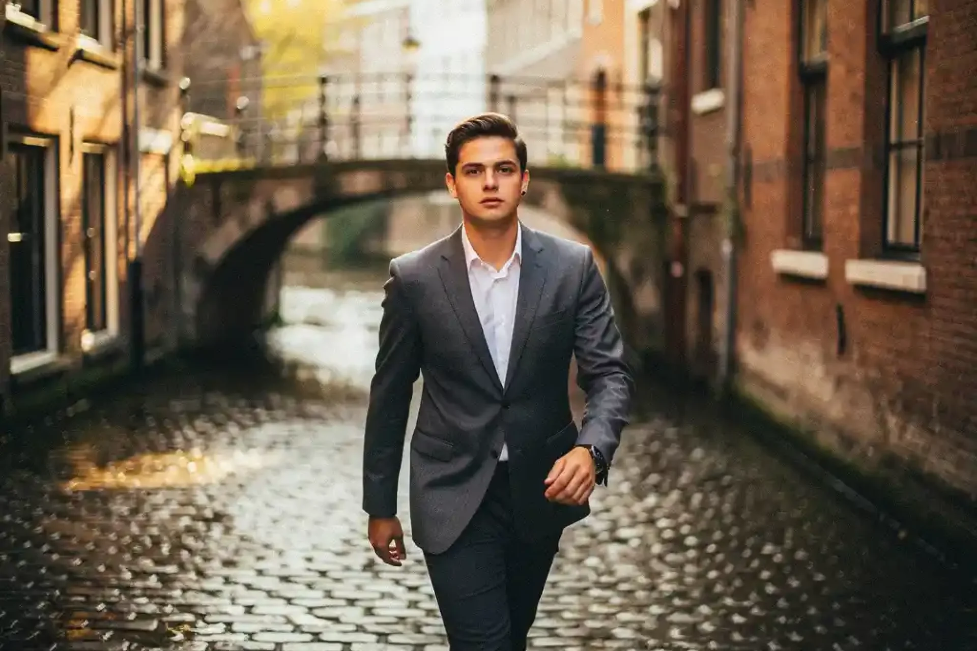 Jonge professional in getailleerd jasje op regenachtige Amsterdamse keiensstraat, historische grachtenpanden in warm middaglicht.