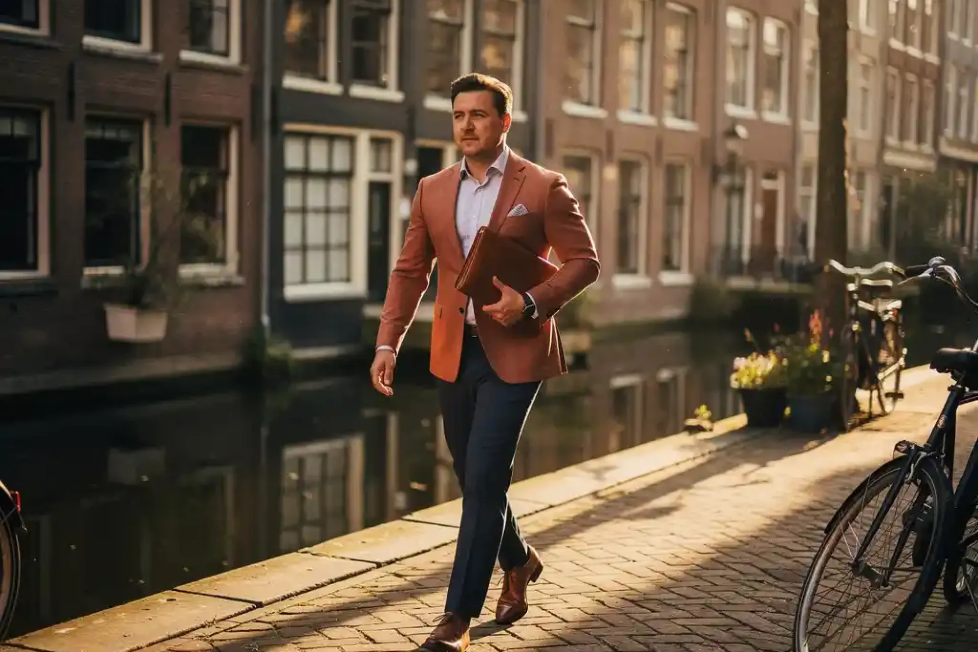 Zelfverzekerde makelaar loopt door smalle Amsterdamse gracht, leren portfolio onder de arm, gouden middagzon op bakstenen gevels en klinkers.