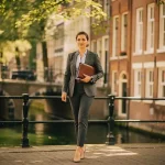 Zelfverzekerde makelaar loopt langs Amsterdams grachtenpand met leren portfolio, historische gevels en boogbrug op de achtergrond.