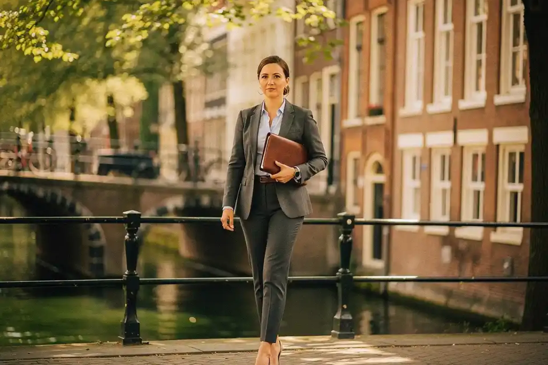 Zelfverzekerde makelaar loopt langs Amsterdams grachtenpand met leren portfolio, historische gevels en boogbrug op de achtergrond.