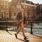 Jonge professional met leren laptoptas loopt zelfverzekerd over een Amsterdamse grachtenbrug, smalle grachtenpanden en waterweg op de achtergrond.