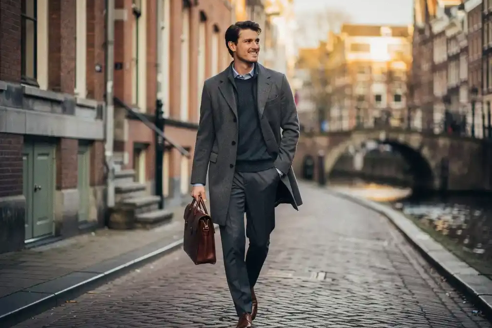 Jonge professional met leren laptoptas loopt over kasseistraatje langs Amsterdamse grachten met historische gevels op de achtergrond.