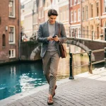 Jonge professional met leren laptoptas en smartphone op smalle Amsterdamse grachtenstraat, historische gevels en boogbrug op achtergrond.