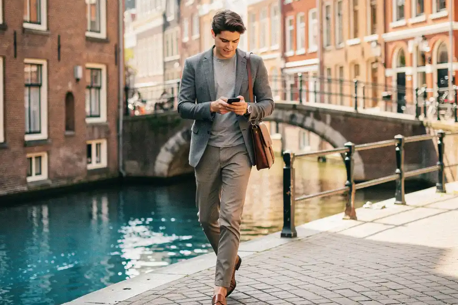 Jonge professional met leren laptoptas en smartphone op smalle Amsterdamse grachtenstraat, historische gevels en boogbrug op achtergrond.