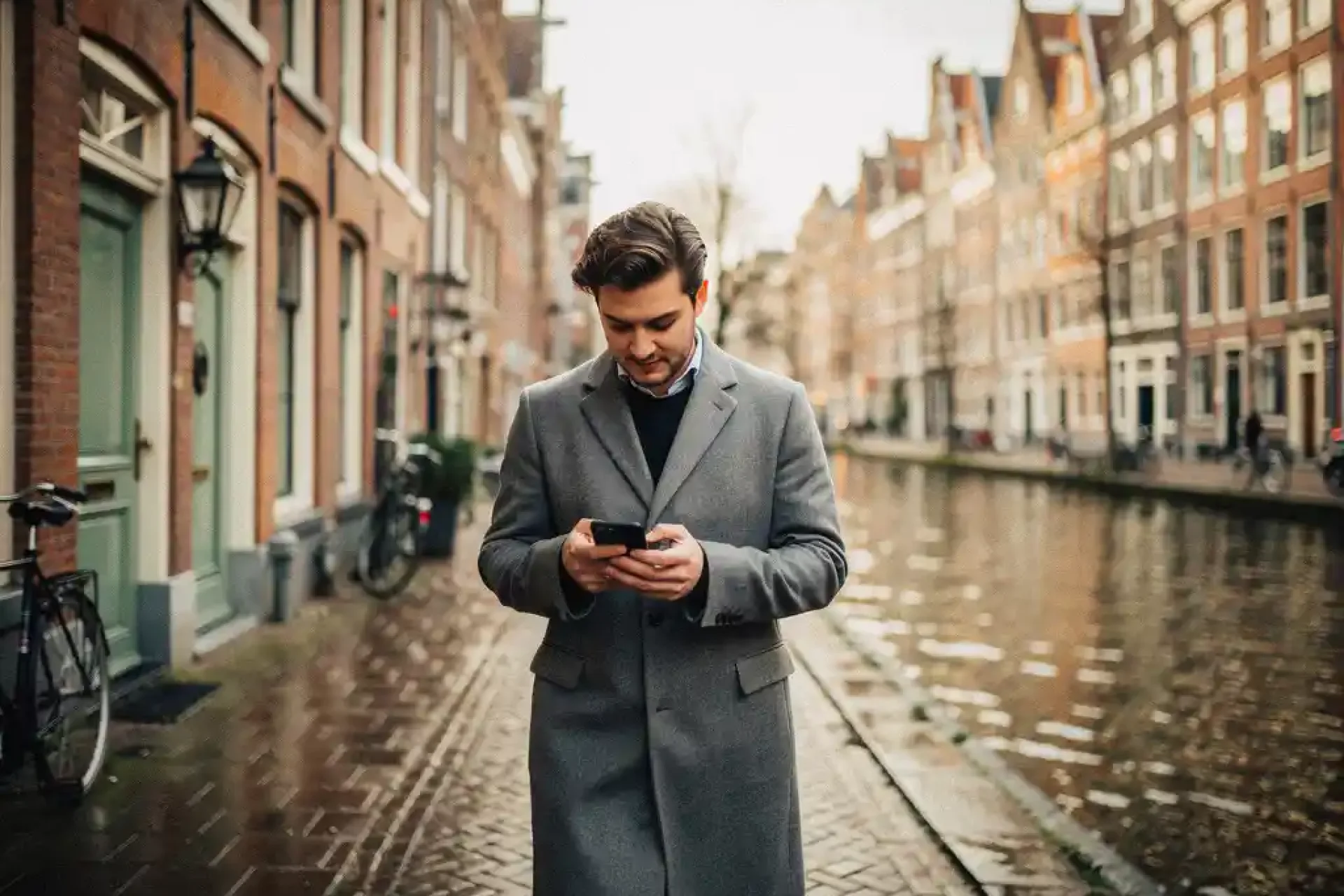 Jonge professional in getailleerde jas loopt door smalle Amsterdamse gracht, checkt notities op telefoon, historische gevels op achtergrond.