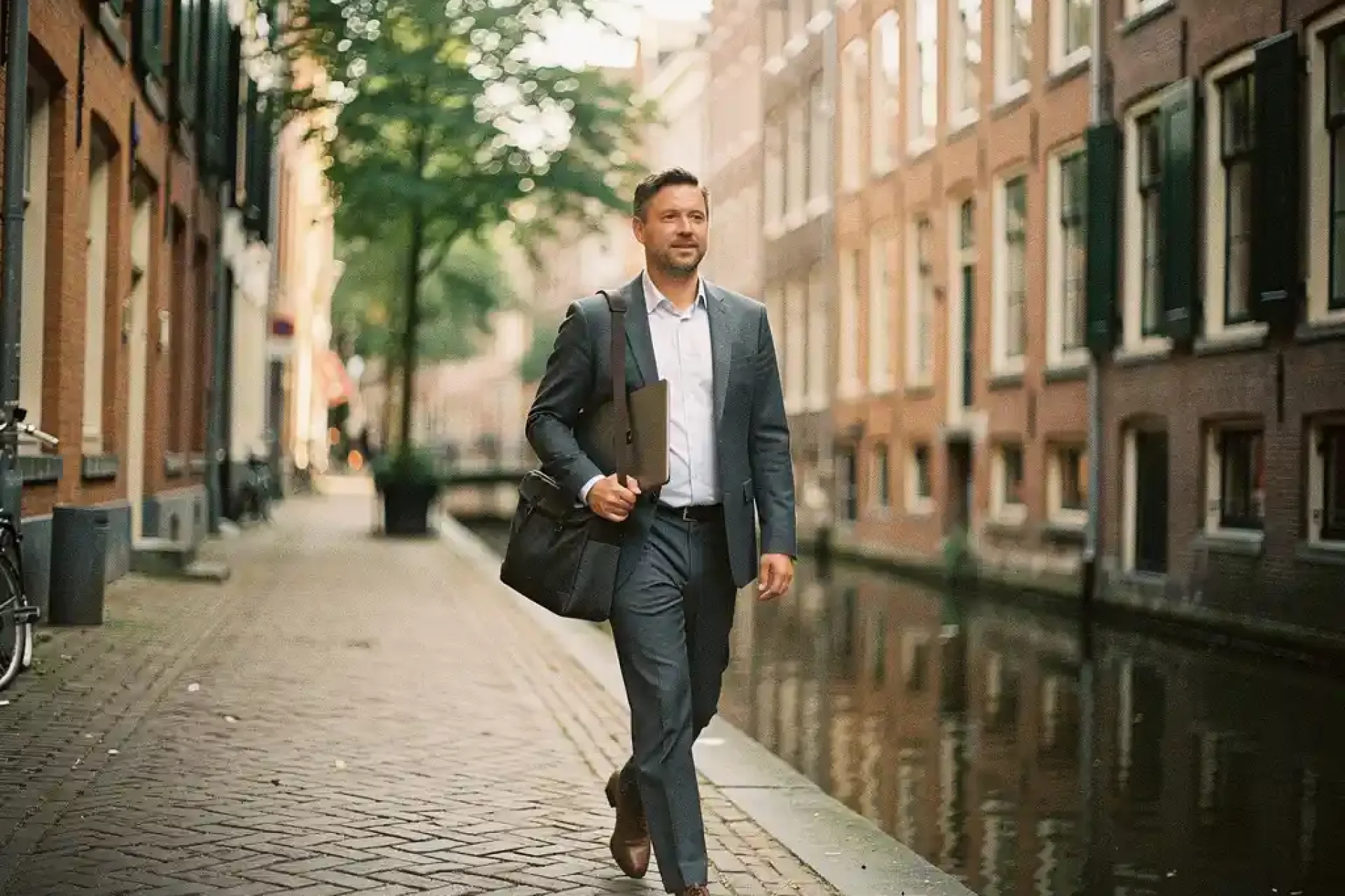 Professional met laptoptas wandelt zelfverzekerd door smal Amsterdams steegje langs historische grachtenpanden en kalm kanalwater.