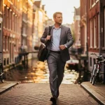 Professional in pak loopt zelfverzekerd door smalle Amsterdamse gracht, laptoptas over schouder, gouden uurtje licht op historische gevels.