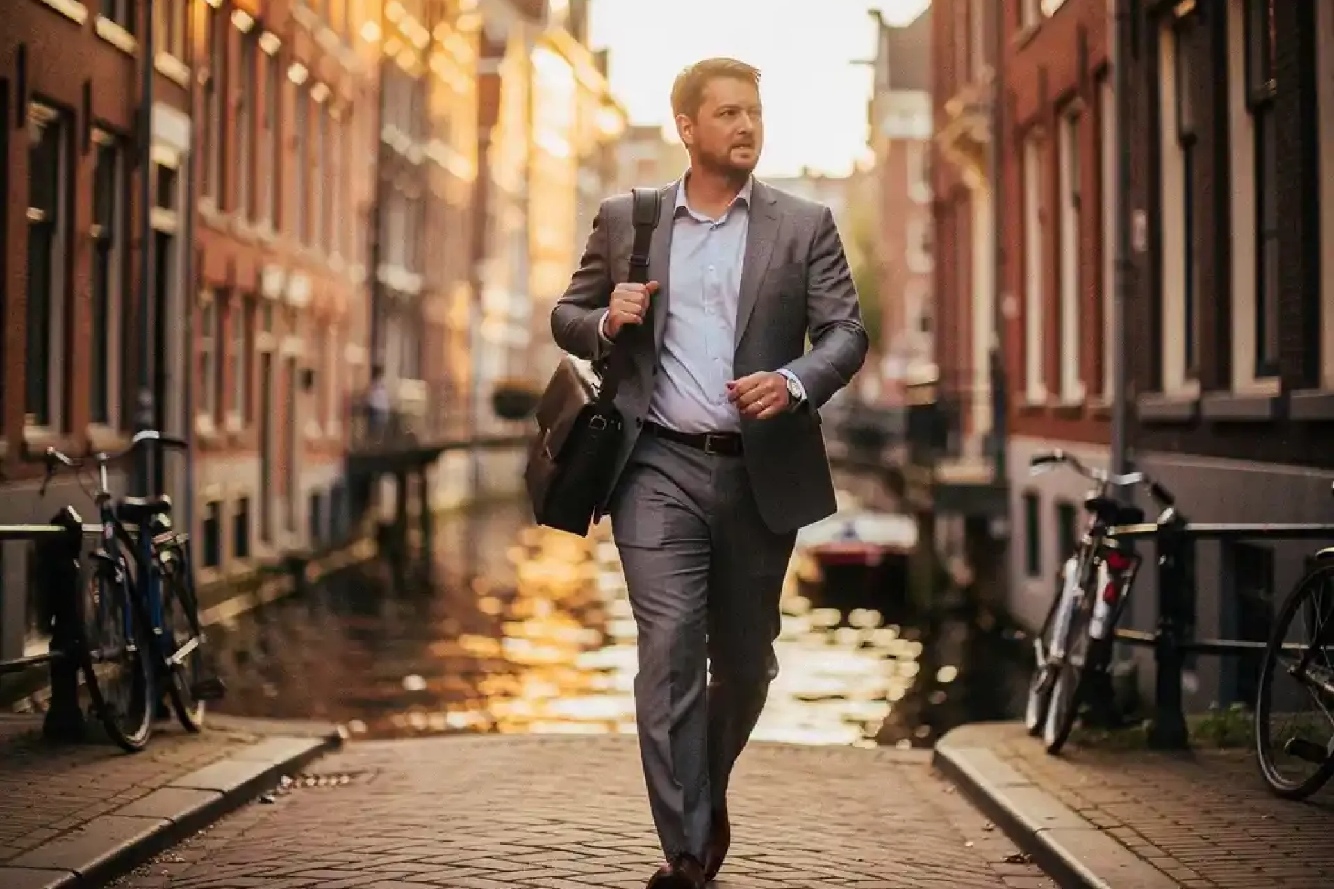 Professional in pak loopt zelfverzekerd door smalle Amsterdamse gracht, laptoptas over schouder, gouden uurtje licht op historische gevels.