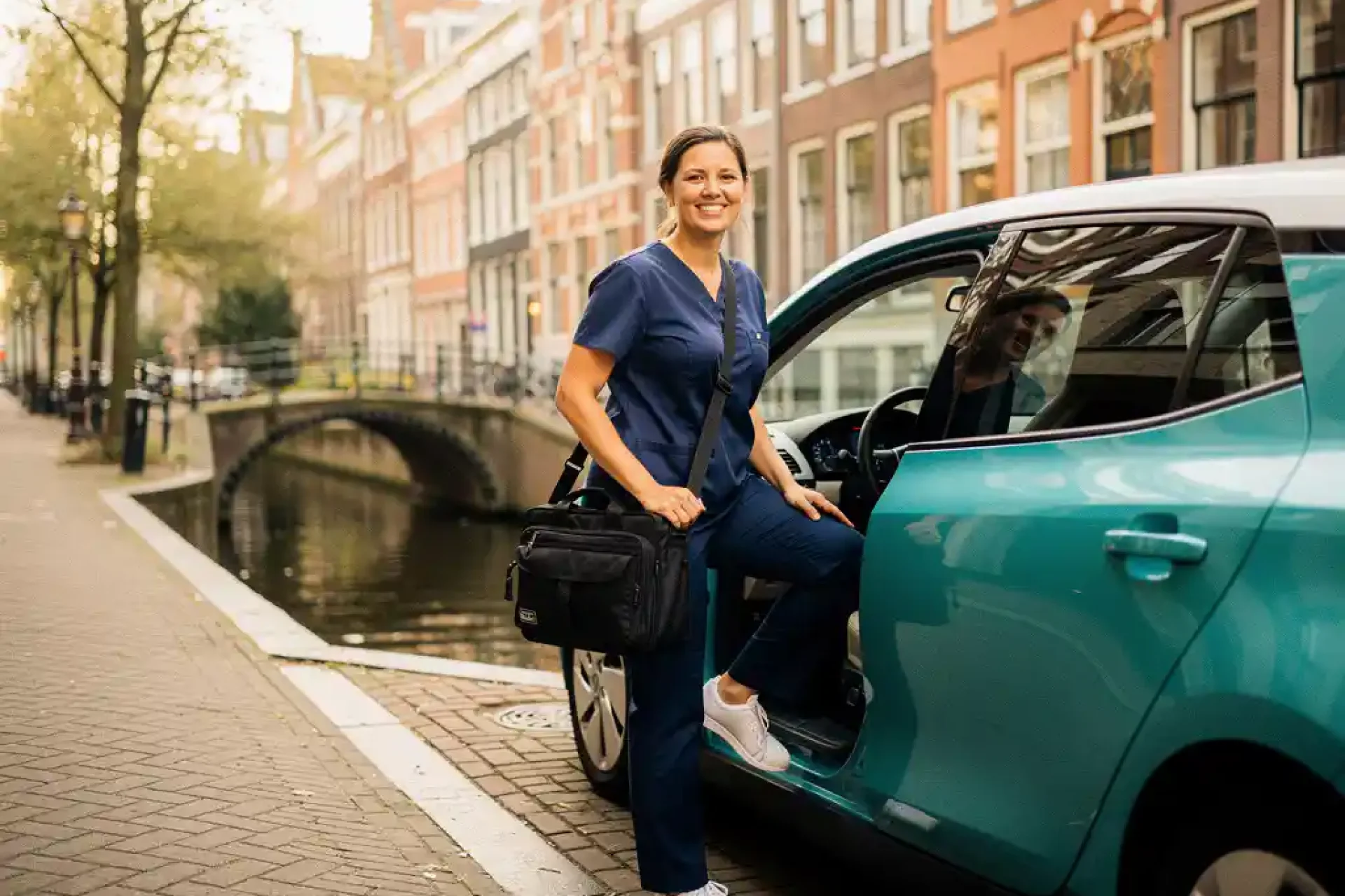 Thuiszorgverpleegkundige in marineblauwe scrubs stapt met medische tas uit elektrische auto op smalle Amsterdamse gracht bij ochtendlicht.