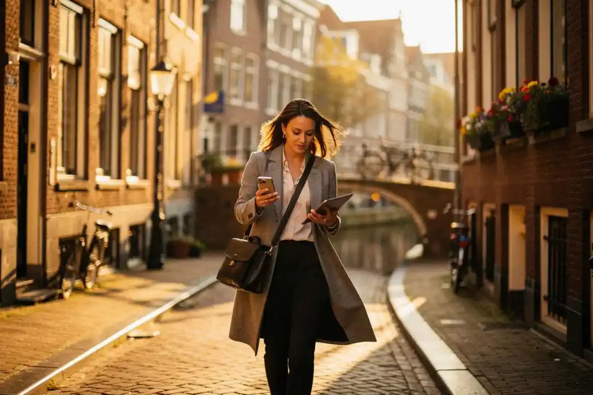 Zelfverzekerde zakenvrouw met laptoptas loopt door smalle Amsterdamse grachtenstraat in gouden middaglicht.