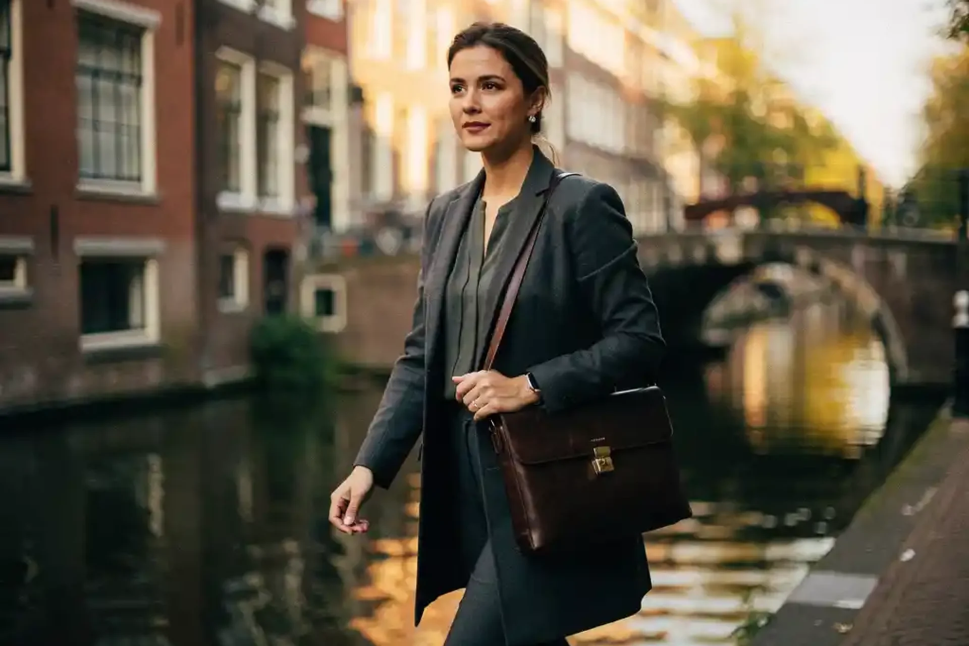 Jonge vrouwelijke professional met leren laptoptas loopt zelfverzekerd langs een Amsterdams gracht, historische gevels op de achtergrond.
