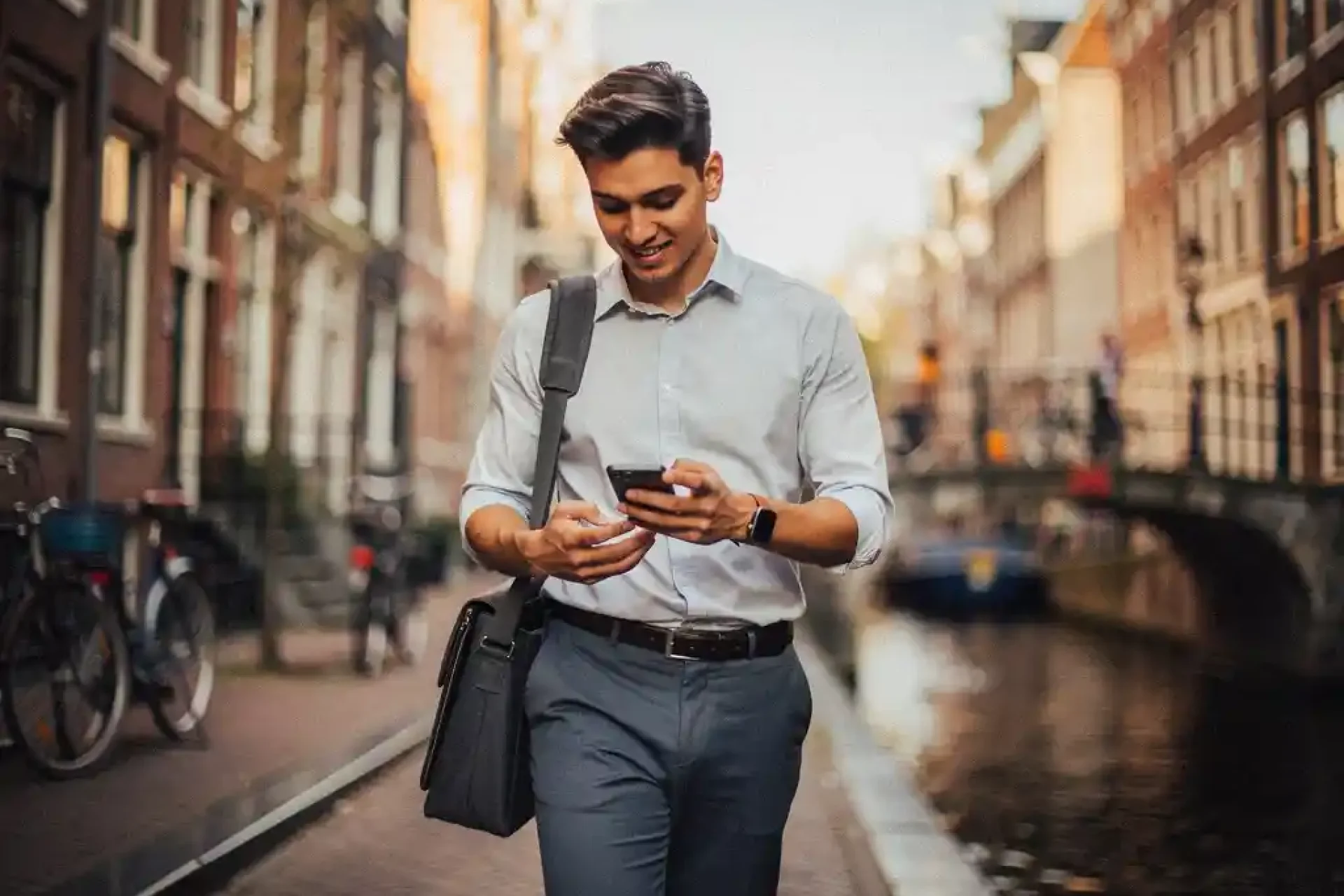 Jonge professional met laptoptas en smartphone op een Amsterdamse grachtenstraat, historische gevels op de achtergrond.