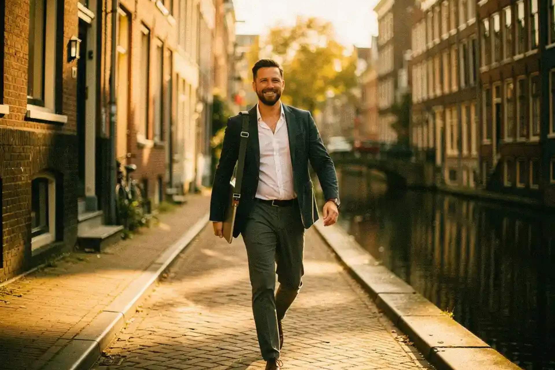 Zelfstandige professional met laptoptas wandelt langs Amsterdams gracht in middagzon, historische bakstenen gevels op achtergrond.